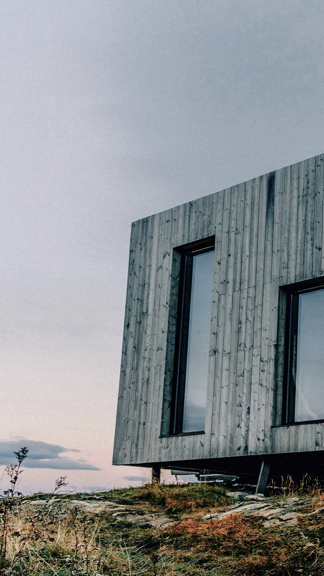 Moderne norsk hytte med grå trekledning og store svarte vinduspartier på eksponert fjelltopp – Natre vinduer