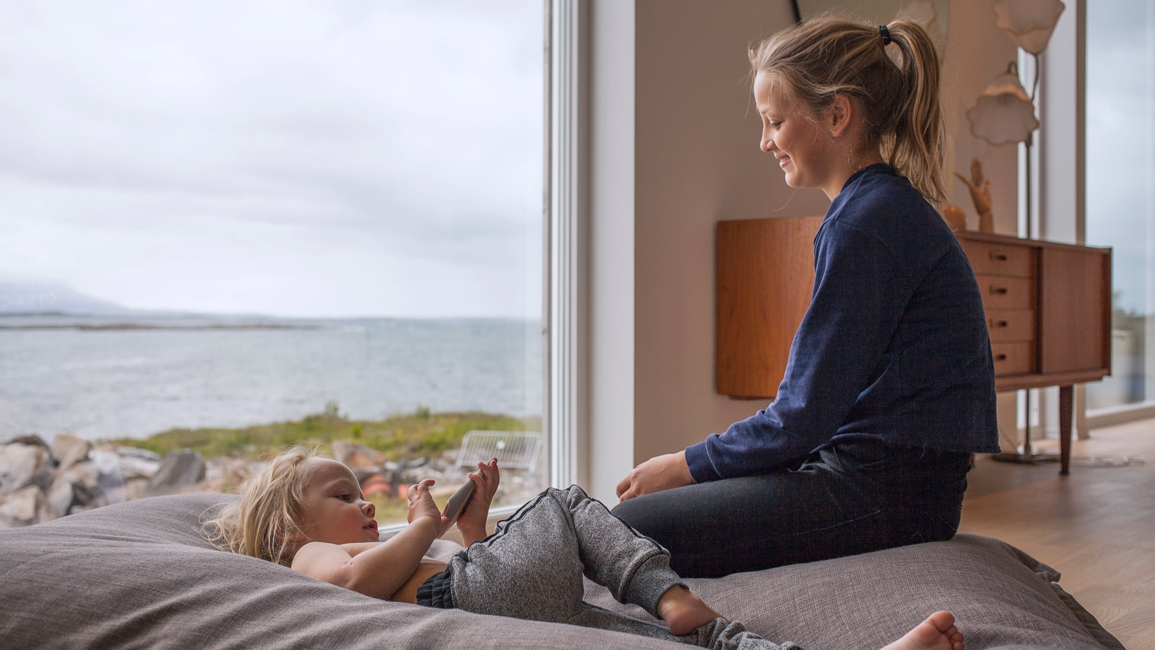 To barn sitter foran stort vindu med havet utenfor
